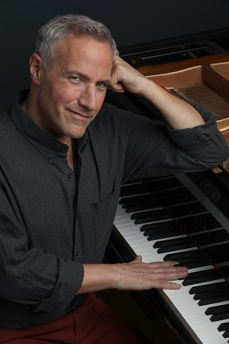 Jim Brickman sitting smiling while leaning against a Grand Piano
