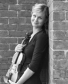Rachael McFarlane, a white woman with hair tied back, leaning against a brick wall and holding her instrument with a smile in black and white