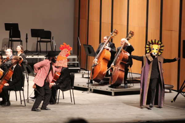 Performers on stage acting as cockerel and lion with costume props to the side of an orchestra 