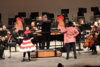 Two performers on stage with chicken and cockerel props standing in front of an orchestra 